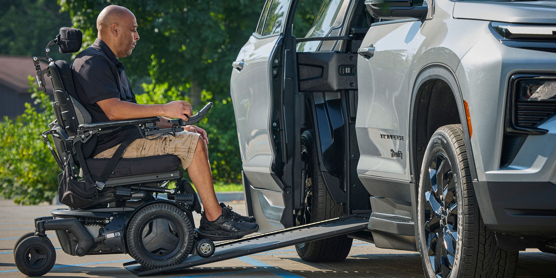 A Person in a Motorized Wheelchair Using an Extended Wheelchair Ramp to Enter a Silver Chevy Traverse SUV Parked Outdoors.